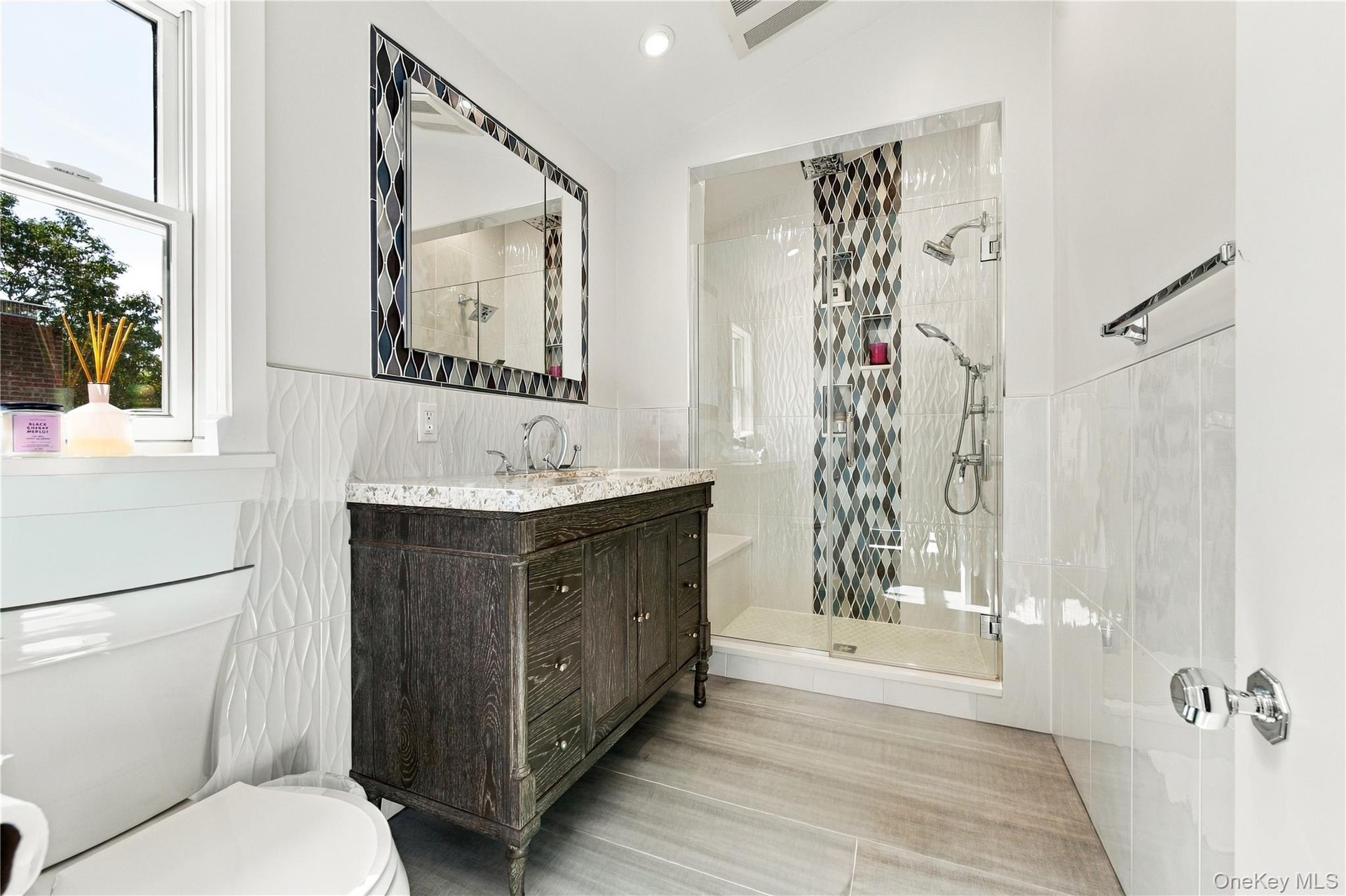 Dark wood-finish vanity with a stone countertop, glass-enclosed shower with mosaic tile inlay, decorative mosaic-framed mirror, and textured wall tile