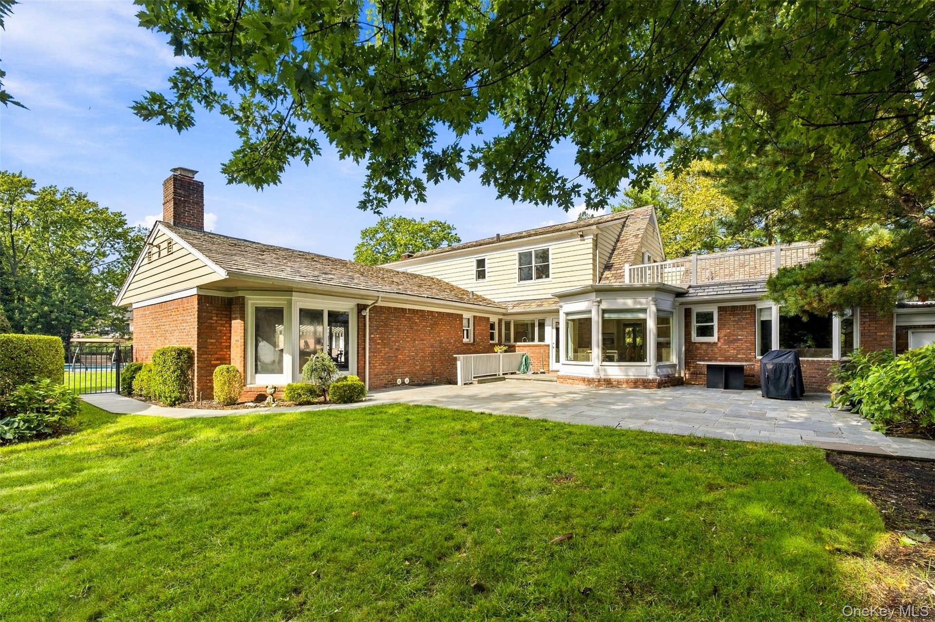 Rear elevation featuring a brick and light-toned horizontal siding exterior, expansive stone patio, masonry chimney, and upper-level railed terrace
