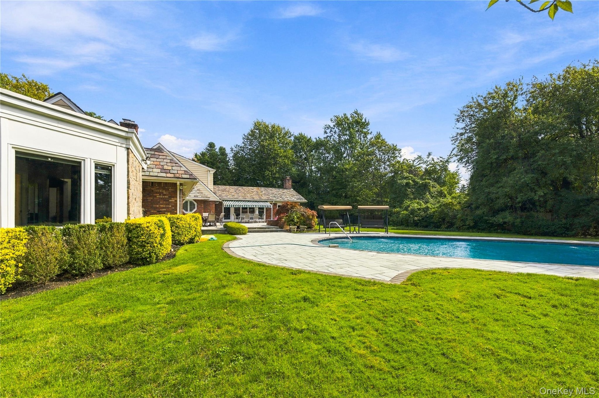 Expansive rear grounds featuring a curved stone pool deck, in-ground swimming pool, manicured lawn, and mature perimeter trees