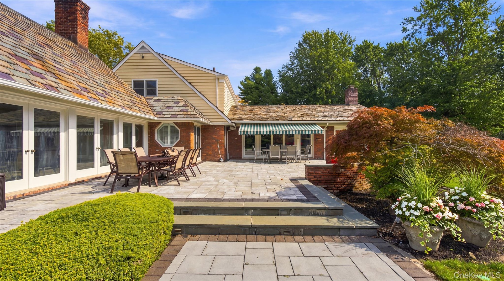 Expansive stone patio featuring a multi-level terrace, brick retaining wall, retractable fabric awning, and slate-style shingle roof