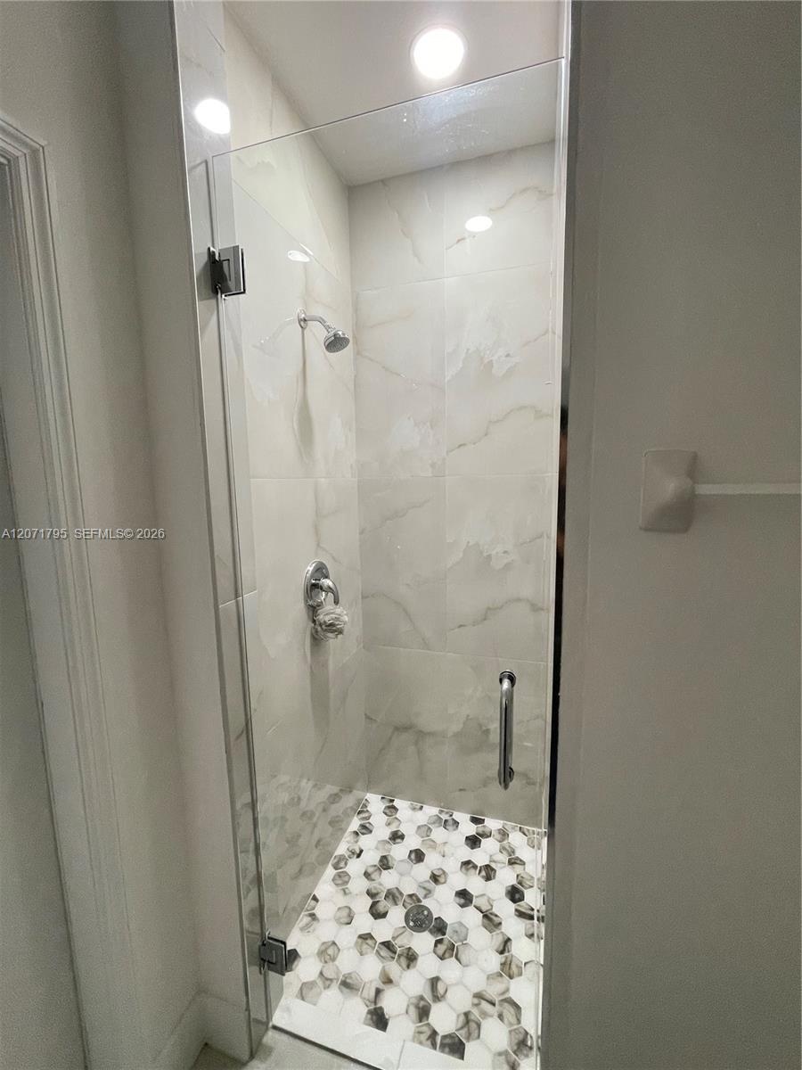 Walk-in shower featuring large-format stone-finish wall tile, a frameless glass door, a metallic hand-held shower head, and decorative hexagonal floor
