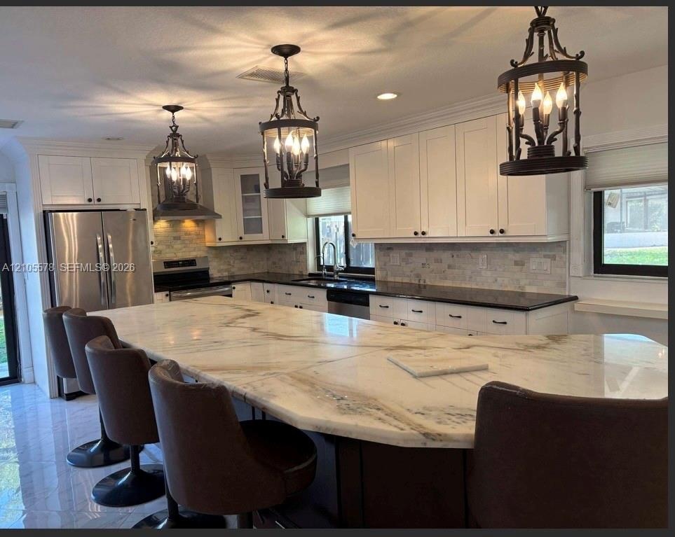 Kitchen featuring a large central island with light-toned natural stone surfacing, white cabinetry, dark countertops, tiled backsplash, and decorative