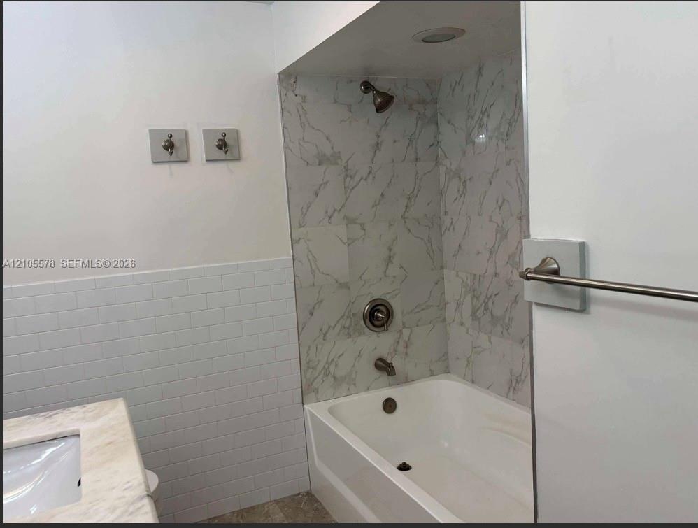Bathtub with stone-finish tile surround, white subway tile wainscoting, integrated countertop vanity, and metal fixtures