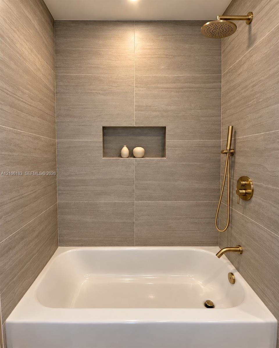 Bathtub with integrated shower featuring large-format tile surround, recessed wall niche, and brushed gold finish fixtures including a rainfall shower