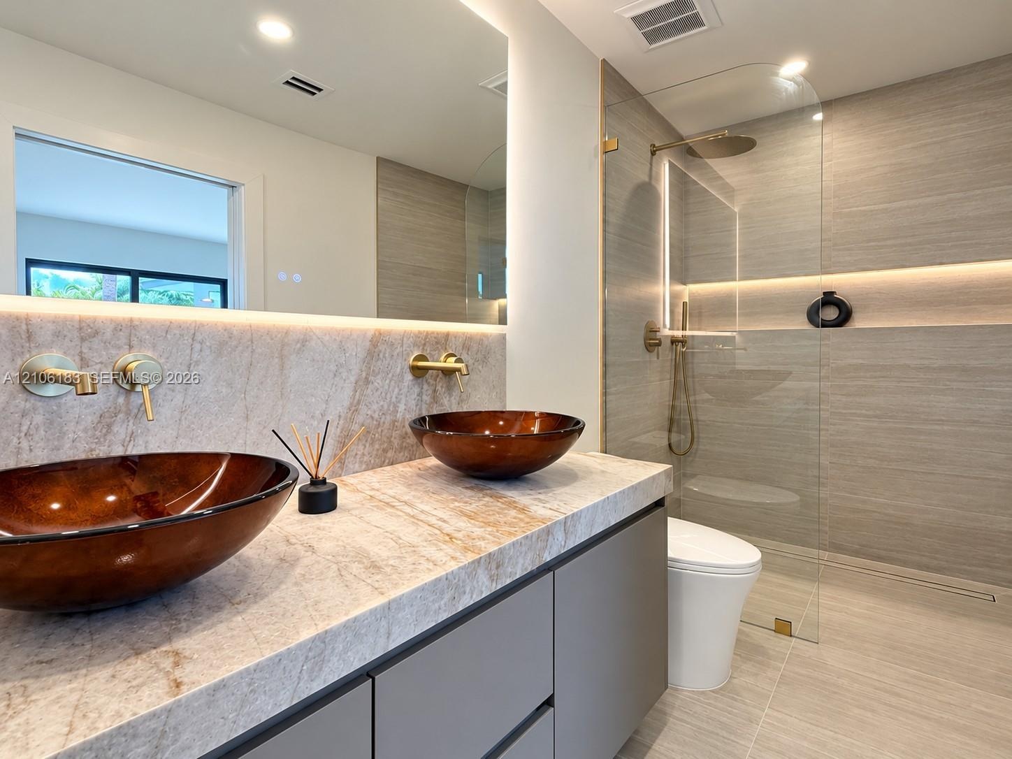 Bathroom vanity featuring a stone countertop, dual glass vessel sinks, wall-mounted gold-tone faucets, and a walk-in shower with floor-to-ceiling tile