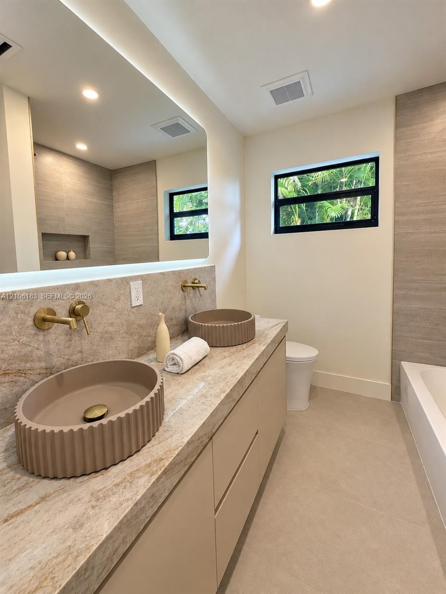 Floating vanity with dual fluted vessel sinks, stone-finish countertop, wall-mounted brass faucets, back-lit vanity mirror, and large-format tile wall