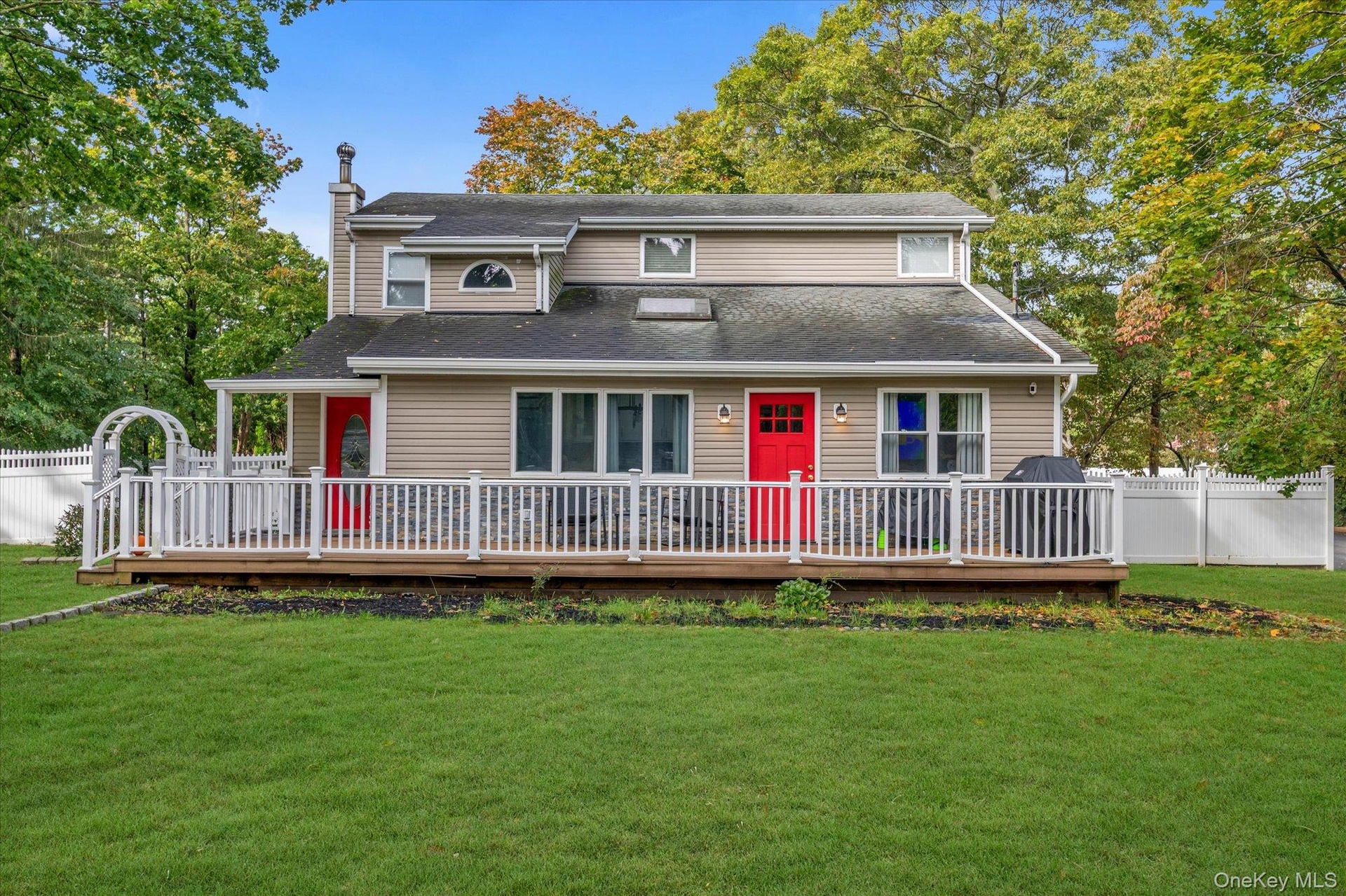 Two-story exterior featuring tan siding, a stone veneer foundation, dual red entry doors, a full-width elevated deck with white railing, and a skyligh
