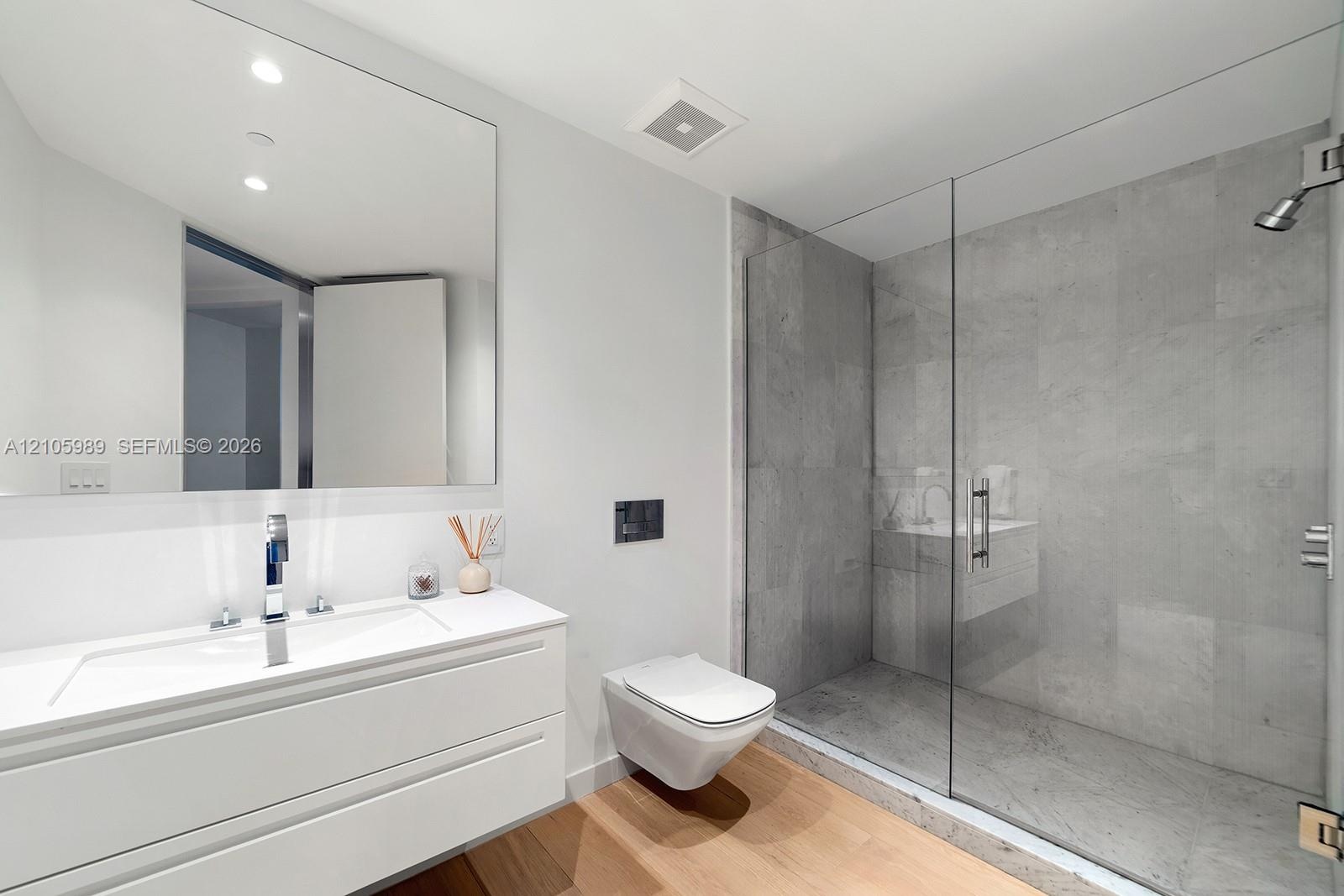 Floating white vanity with a rectangular sink, wall-mounted toilet, walk-in shower with a glass enclosure and stone-tile surround, and wood-finish flo