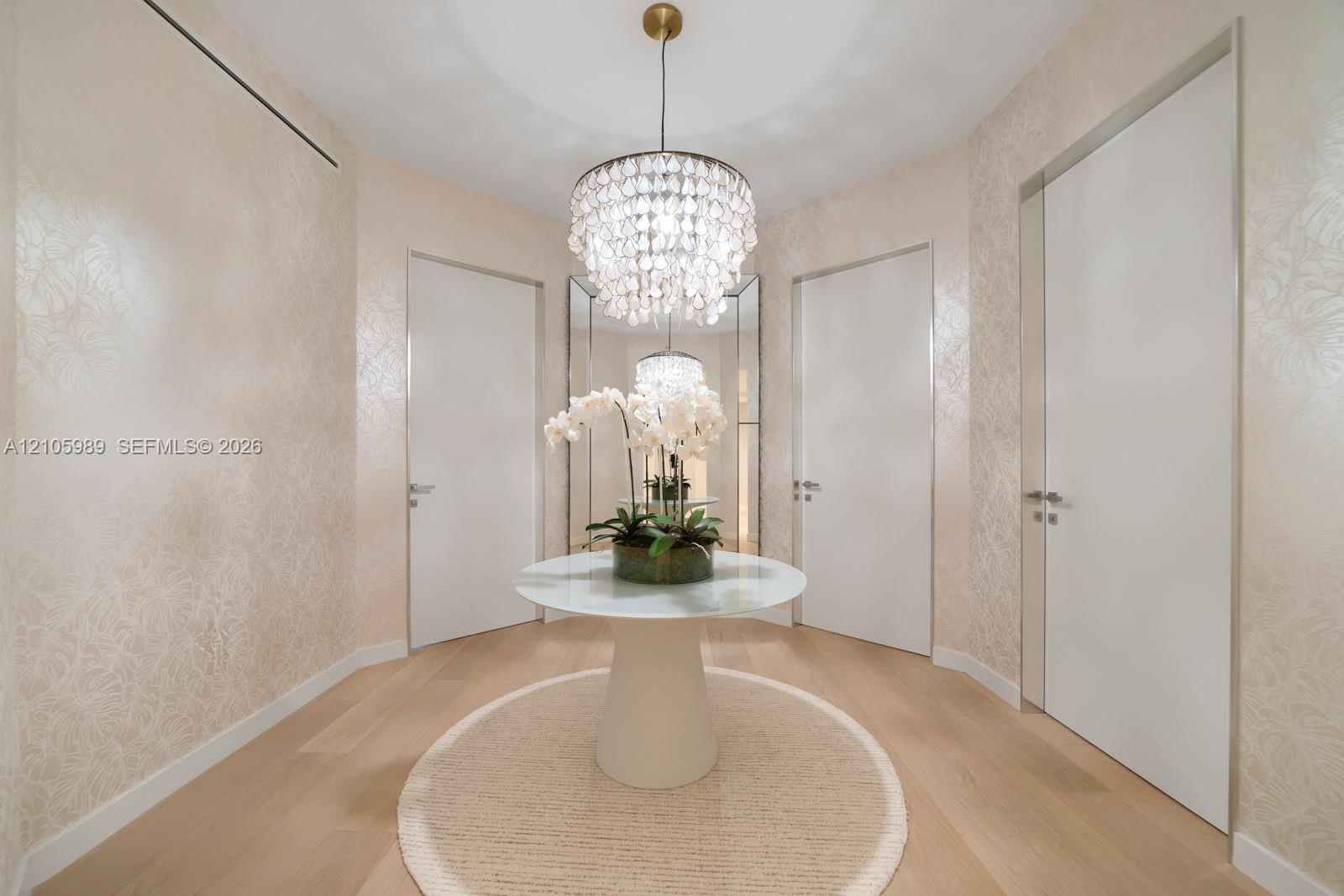Foyer featuring light-toned wood-finish flooring, textured wall coverings, a tiered crystal chandelier, full-height flush doors with metallic casing, 