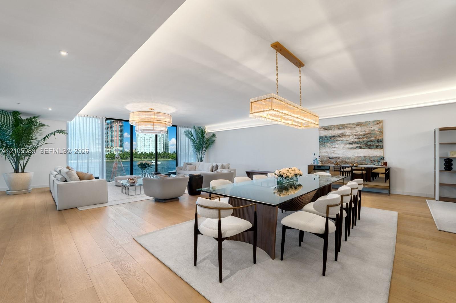 Open-concept living space featuring wood-finish flooring, recessed perimeter ceiling lighting, floor-to-ceiling windows, and dual statement chandelier