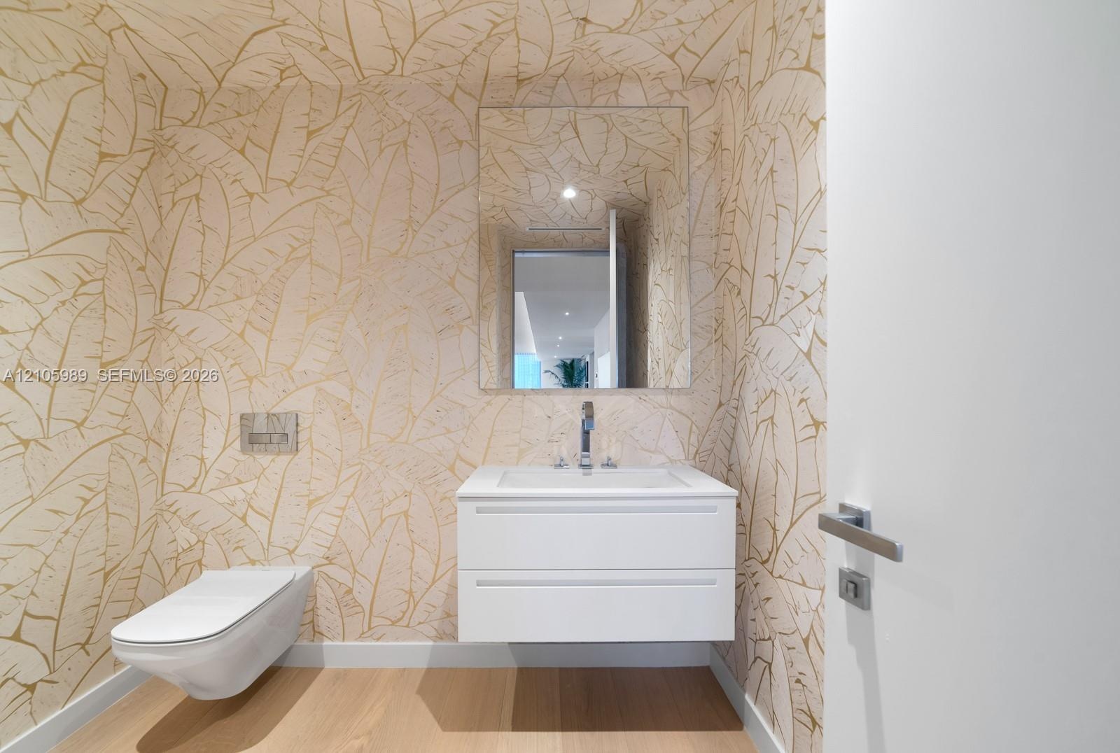 Floating white vanity with a rectangular sink, wall-mounted dual-flush toilet, large frameless mirror, wood-finish flooring, and metallic patterned wa