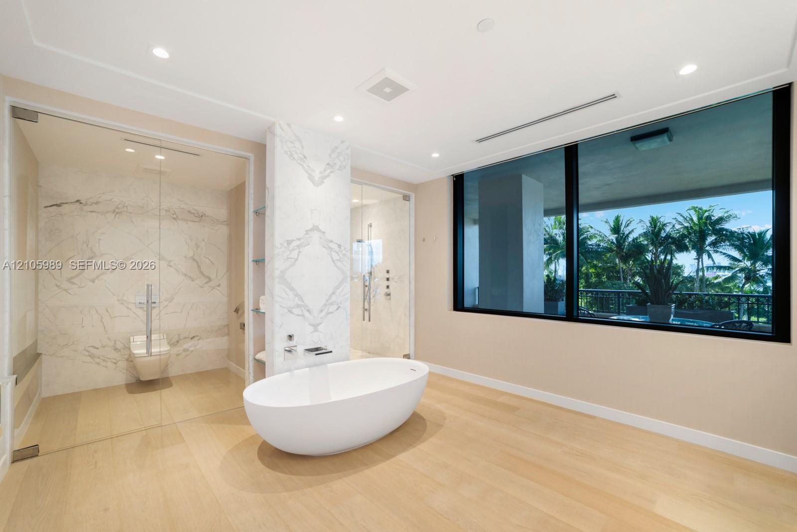 Freestanding soaking tub, stone-clad feature wall, glass-enclosed shower, wood-finish flooring, and expansive window with exterior greenery