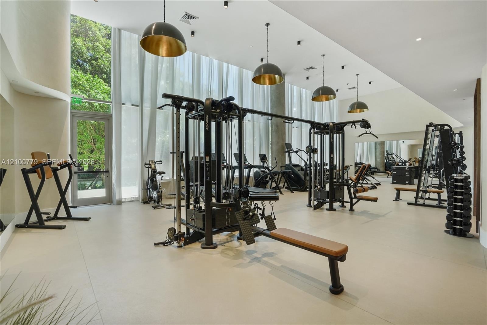 Fitness space featuring floor-to-ceiling windows, neutral tile flooring, recessed ceiling lighting, and suspended pendant fixtures with metallic inter