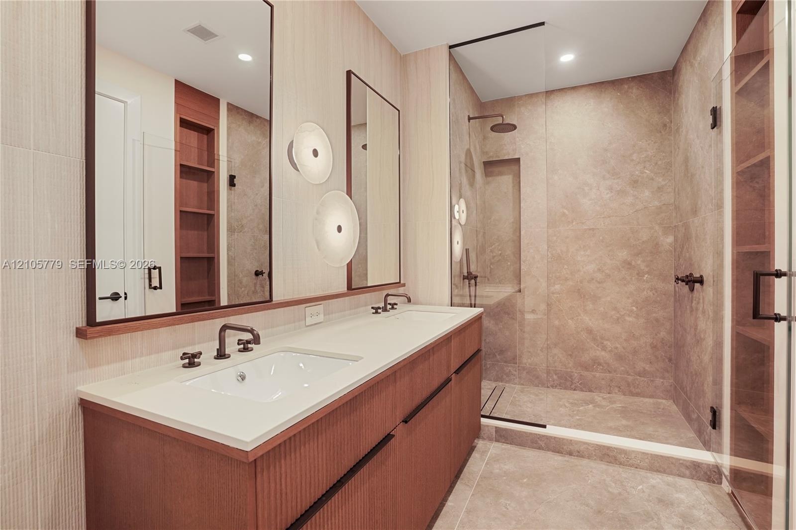 Double vanity with a composite countertop, fluted wood-finish cabinetry, walk-in shower with stone-tile surround, glass enclosure, and wall-mounted pl