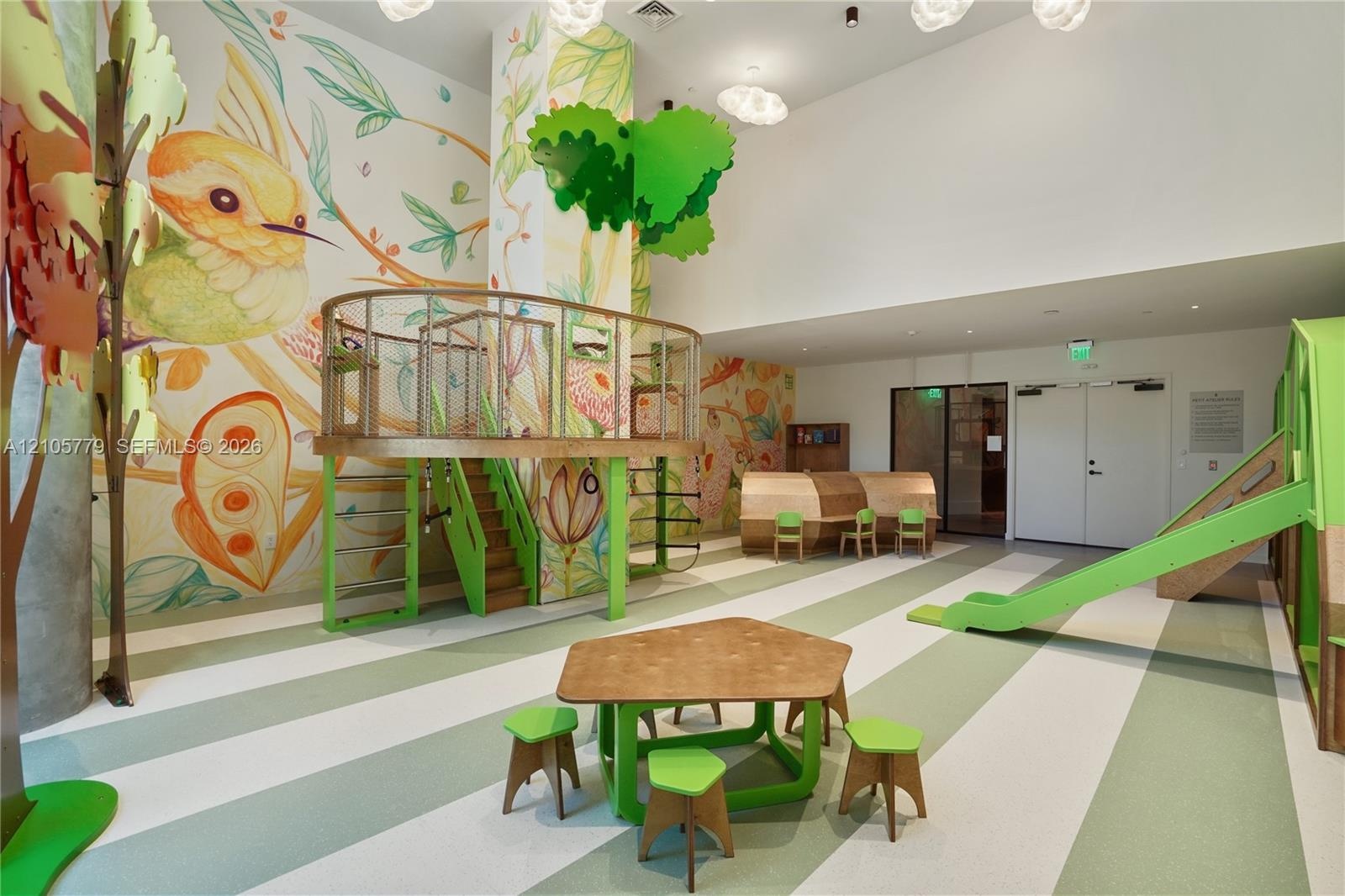 Indoor recreational space featuring a high ceiling, full-wall nature-themed mural, built-in wooden play structure with platform, integrated slide, and