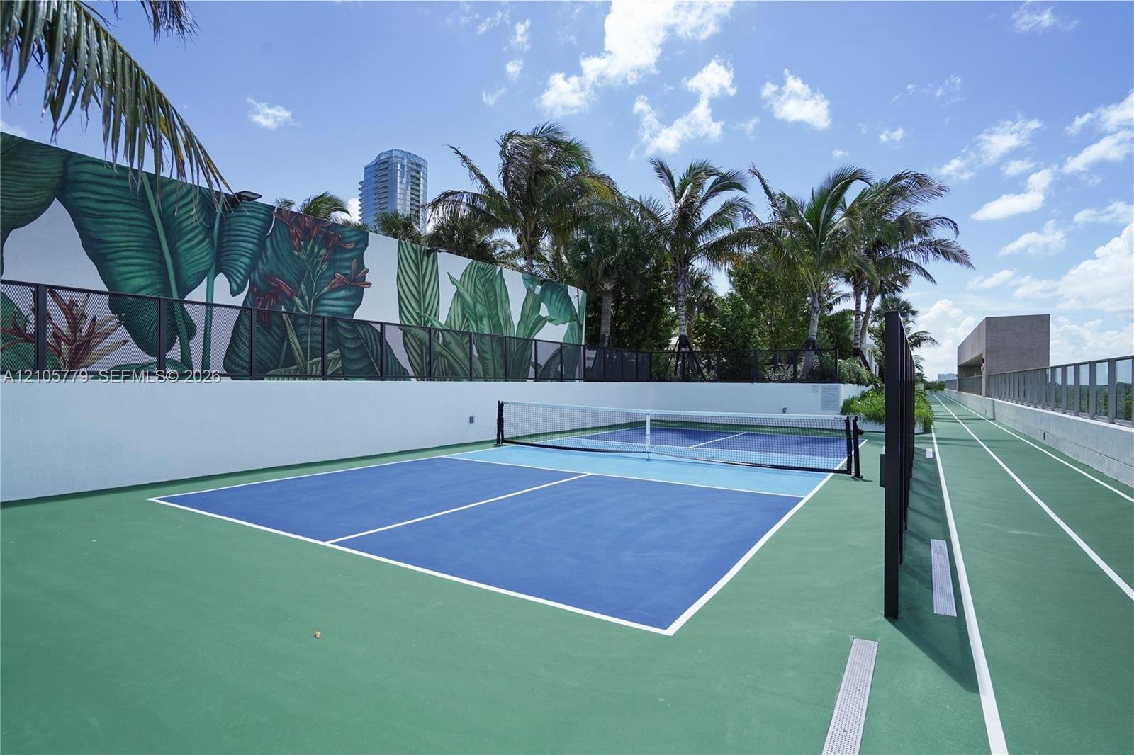 Rooftop pickleball court featuring a multi-toned blue and green athletic surface, perimeter fencing, and a decorative mural wall