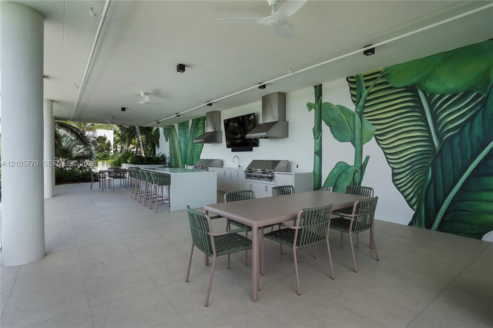 Covered outdoor kitchen featuring a large stone-finish island with integrated seating, built-in stainless steel appliances, dual vent hoods, and overs