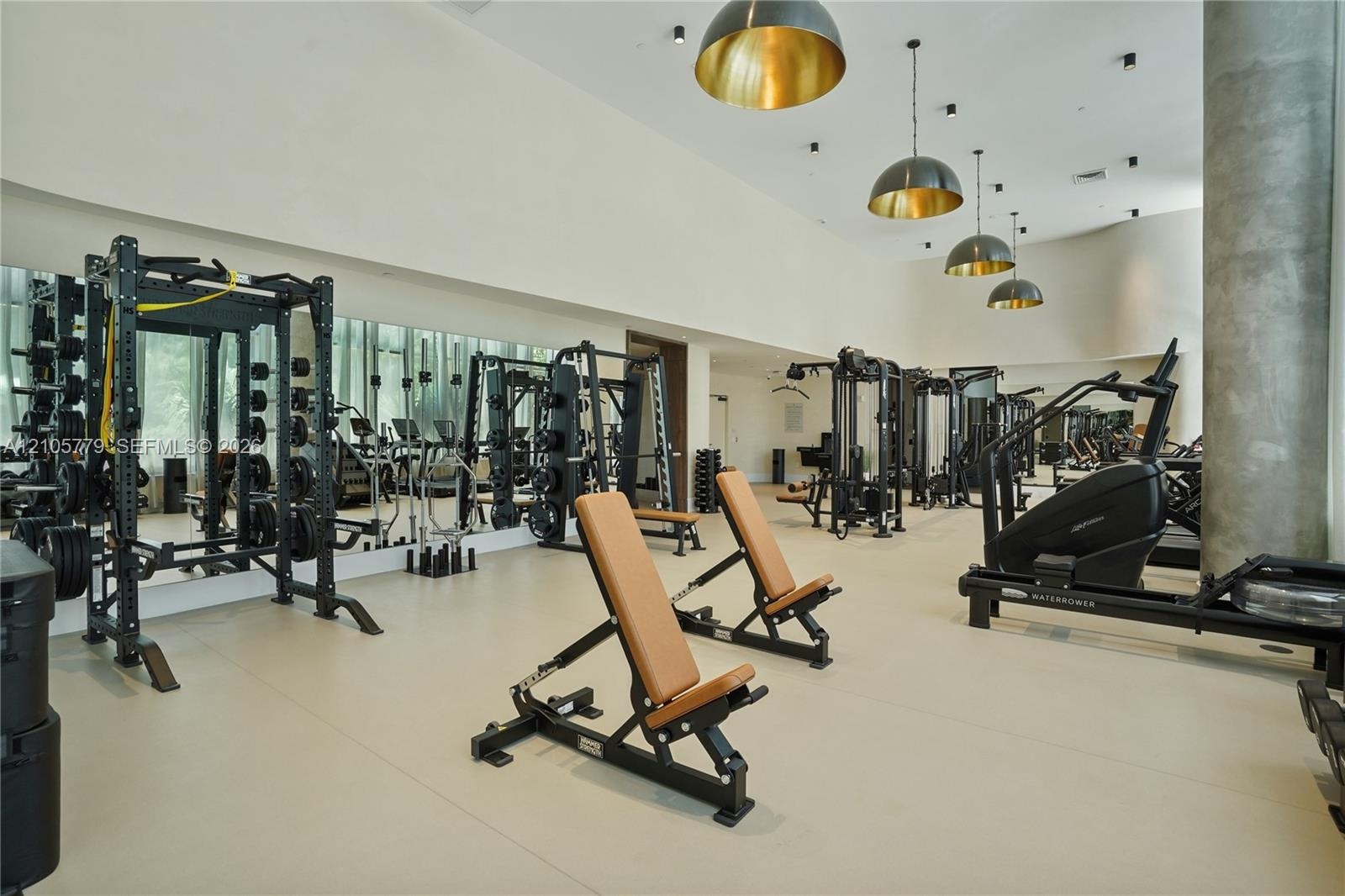 High-ceiling fitness center featuring expansive wall-to-wall mirrors, industrial-style dome pendant lighting, and concrete-finish support columns