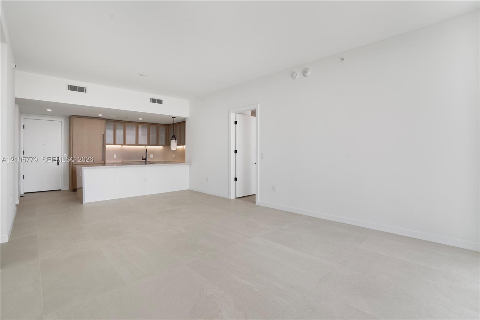 Open-concept interior with large-format neutral tile flooring, minimalist kitchen featuring light wood-tone cabinetry, stone-surface breakfast bar, an