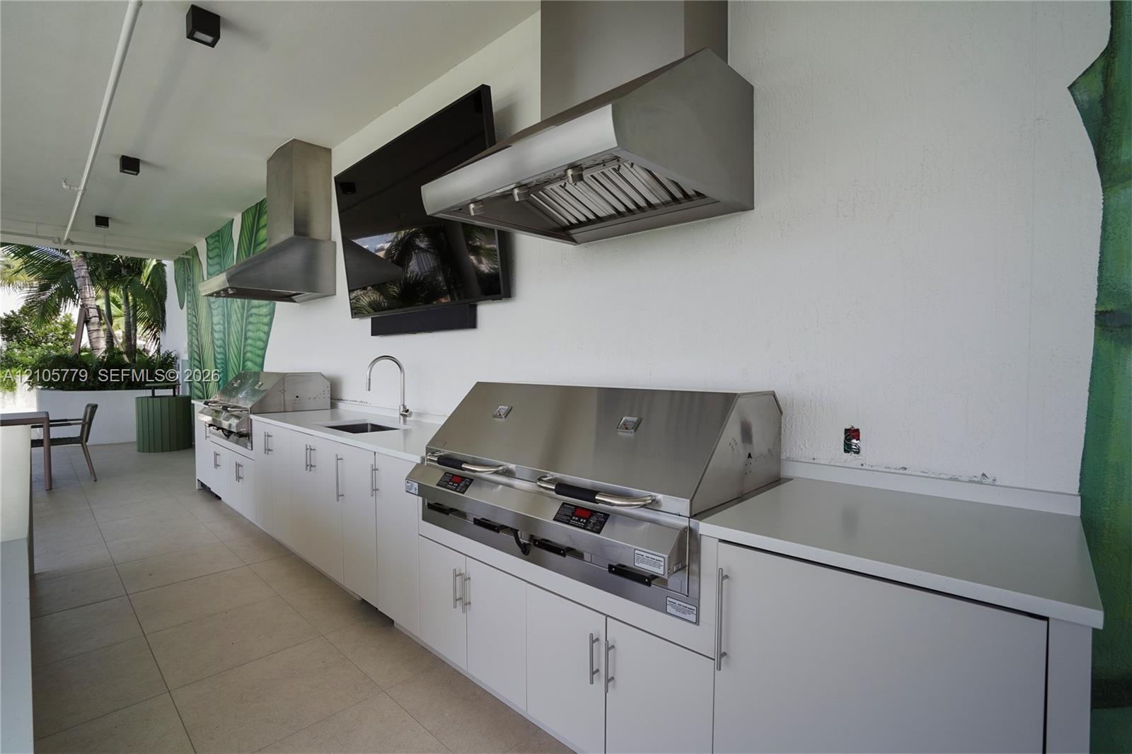 Outdoor kitchen featuring dual stainless steel ventilation hoods, built-in stainless steel grill, integrated sink with gooseneck faucet, and white cab