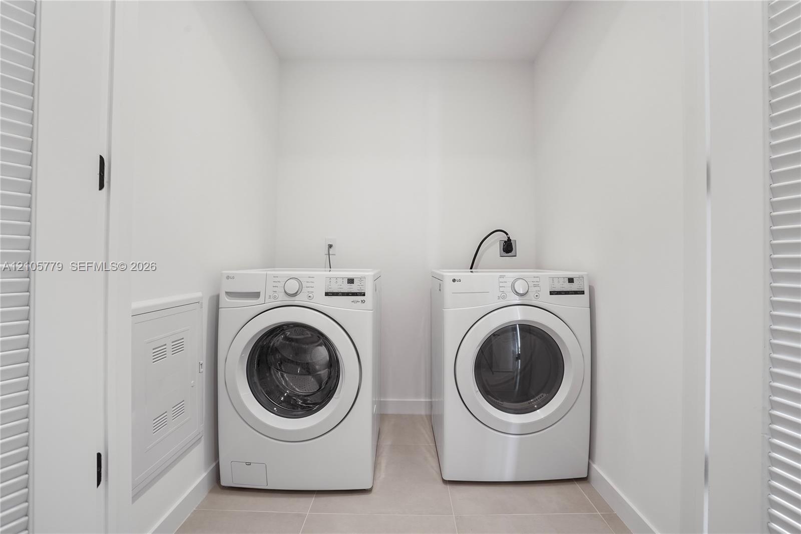 Laundry space featuring white front-loading appliances, neutral tile flooring, and louvered privacy doors