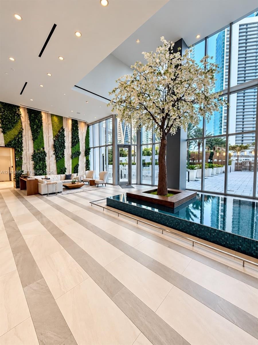 Double-height lobby featuring floor-to-ceiling glass walls, an indoor reflecting pool, a central ornamental tree, and a vertical green wall installati