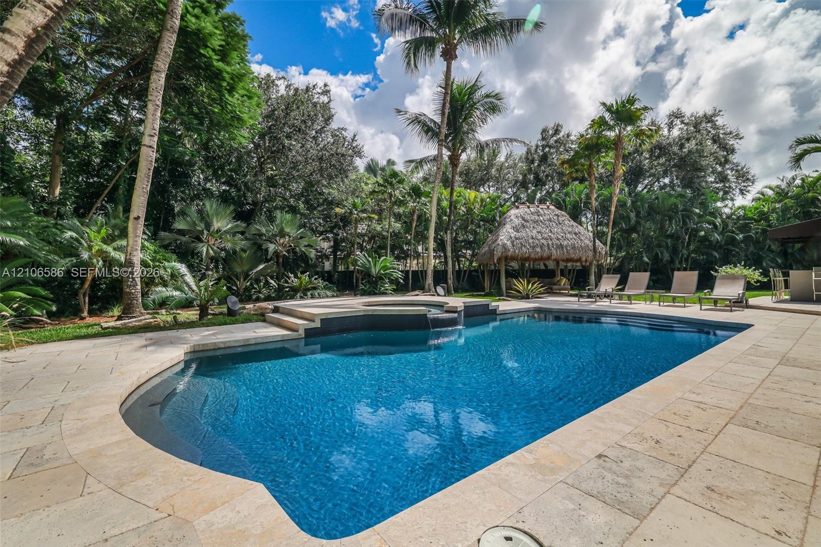 In-ground swimming pool with an integrated elevated spa, stone paver decking, and a detached thatched-roof structure surrounded by mature palms and la