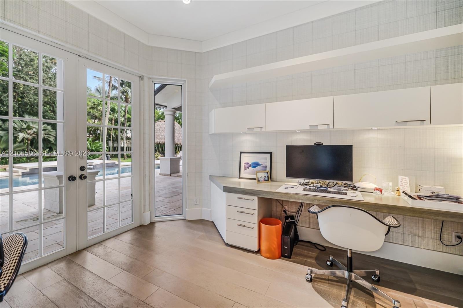 Custom built-in desk with integrated storage cabinetry, wood-finish flooring, and white paneled walls featuring multi-pane glass doors
