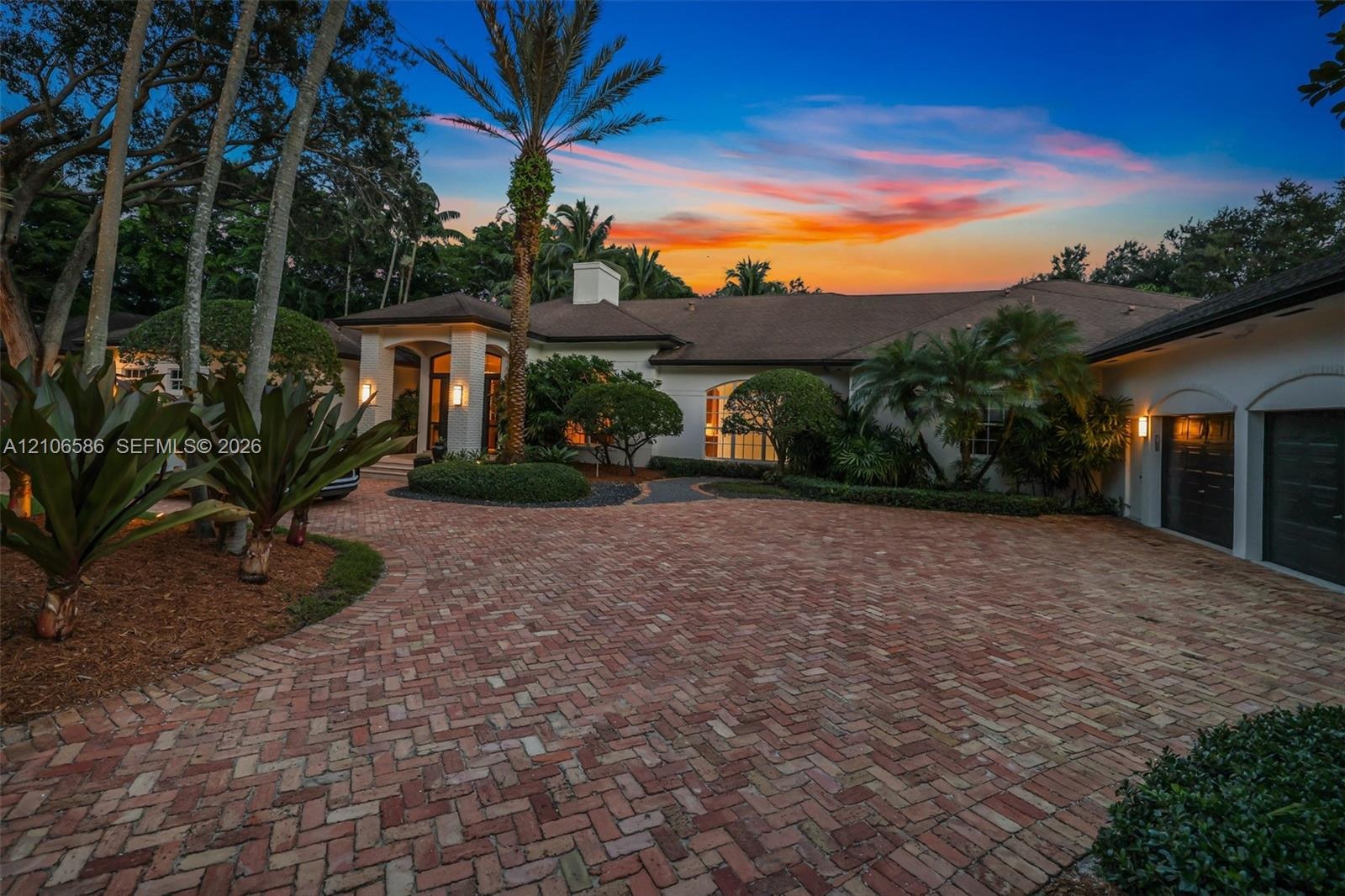 Stucco exterior featuring a herringbone-pattern brick paver driveway, covered entryway, multi-car garage, and mature landscaping