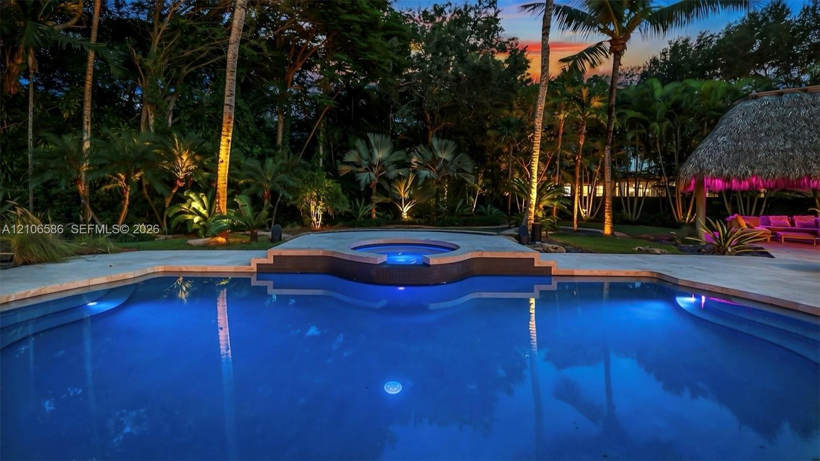In-ground swimming pool with integrated elevated spa, stone-paver decking, thatch-roof cabana, and mature perimeter landscaping