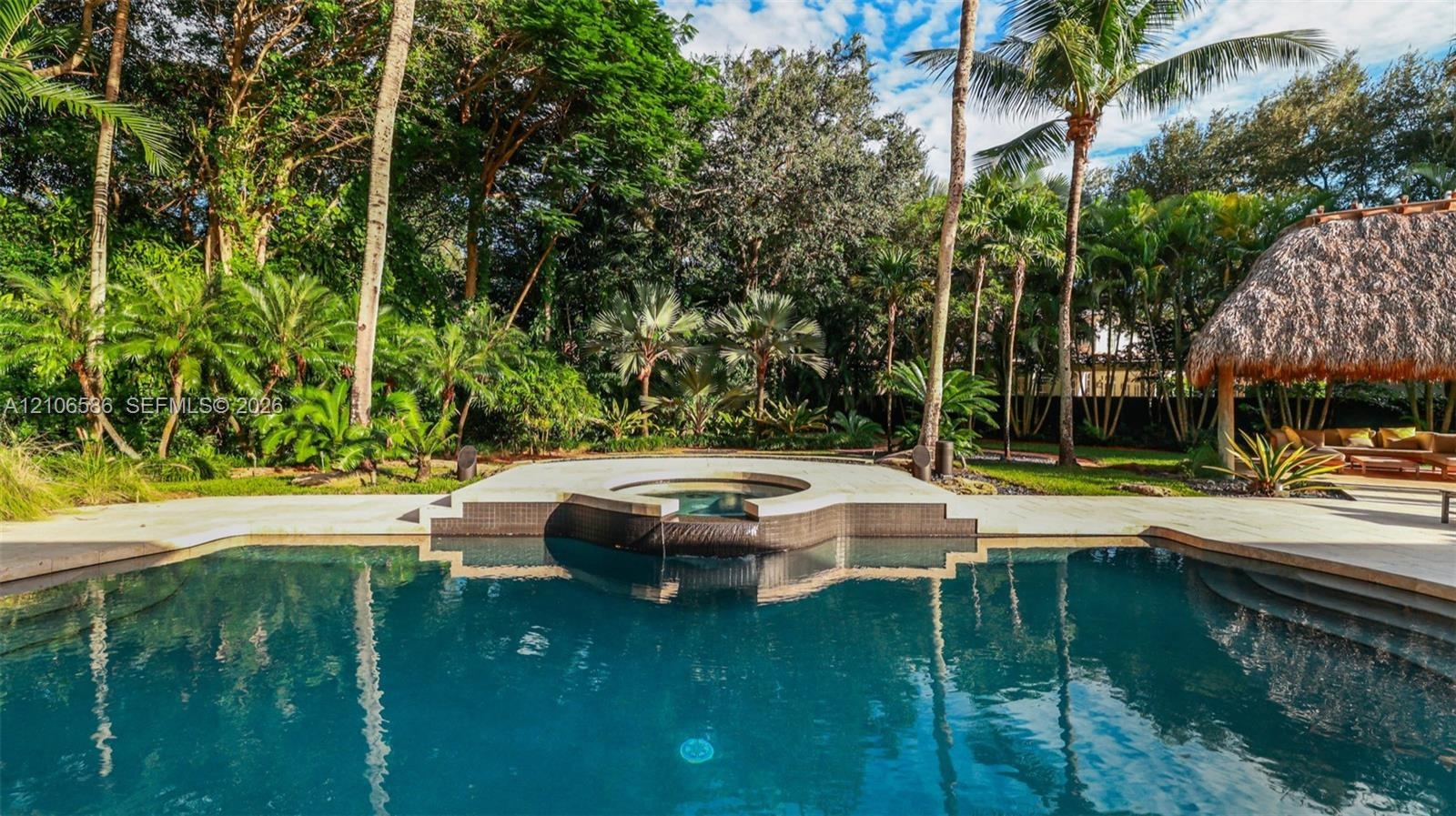 In-ground swimming pool featuring a tiered spillover spa, stone-tile surround, and a thatched-roof palapa set against a landscaped backdrop of mature 