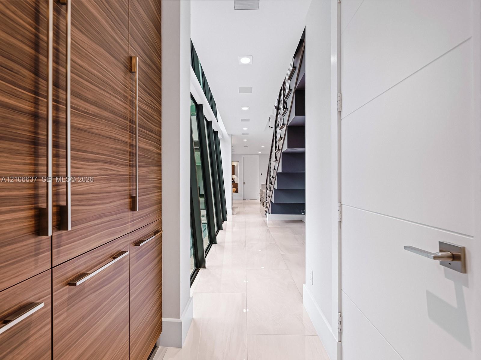 Hallway featuring wood-finish cabinetry with vertical hardware, large-format tile flooring, recessed ceiling lighting, and a dark-toned staircase with