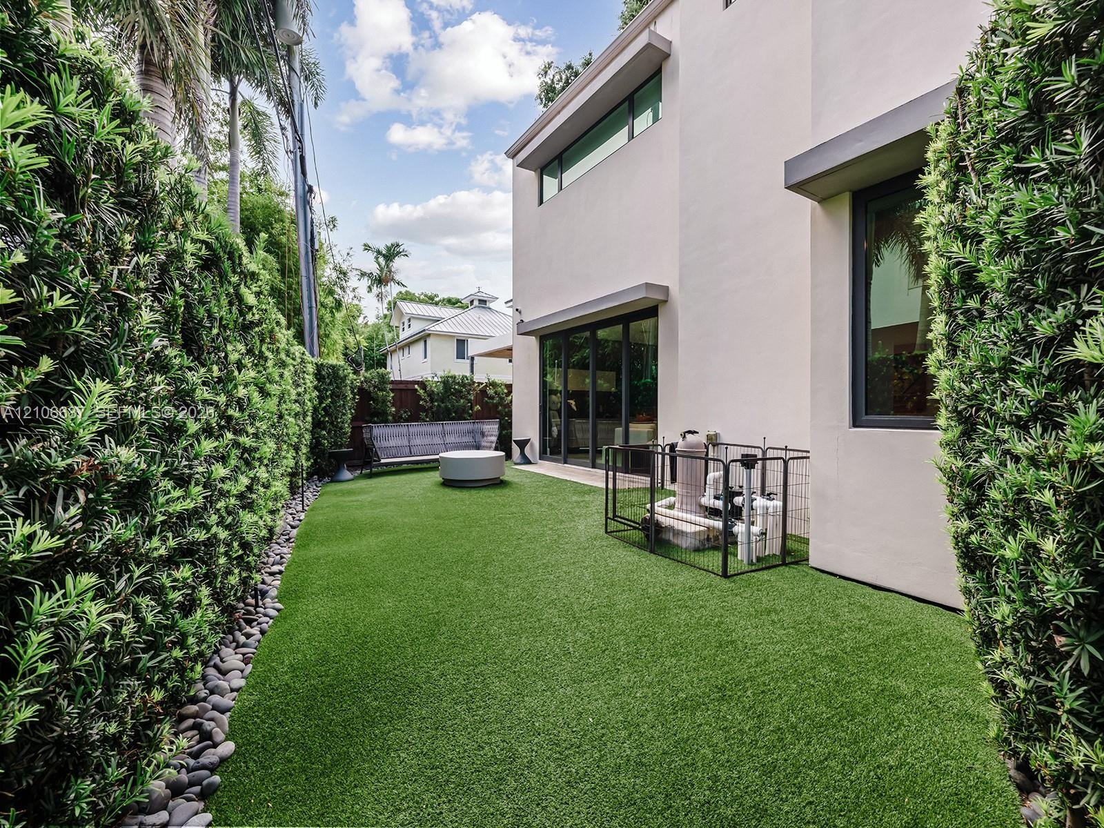 Modern exterior featuring synthetic turf, structured evergreen hedges, smooth stucco siding, large-format sliding glass doors, and dark-framed windows