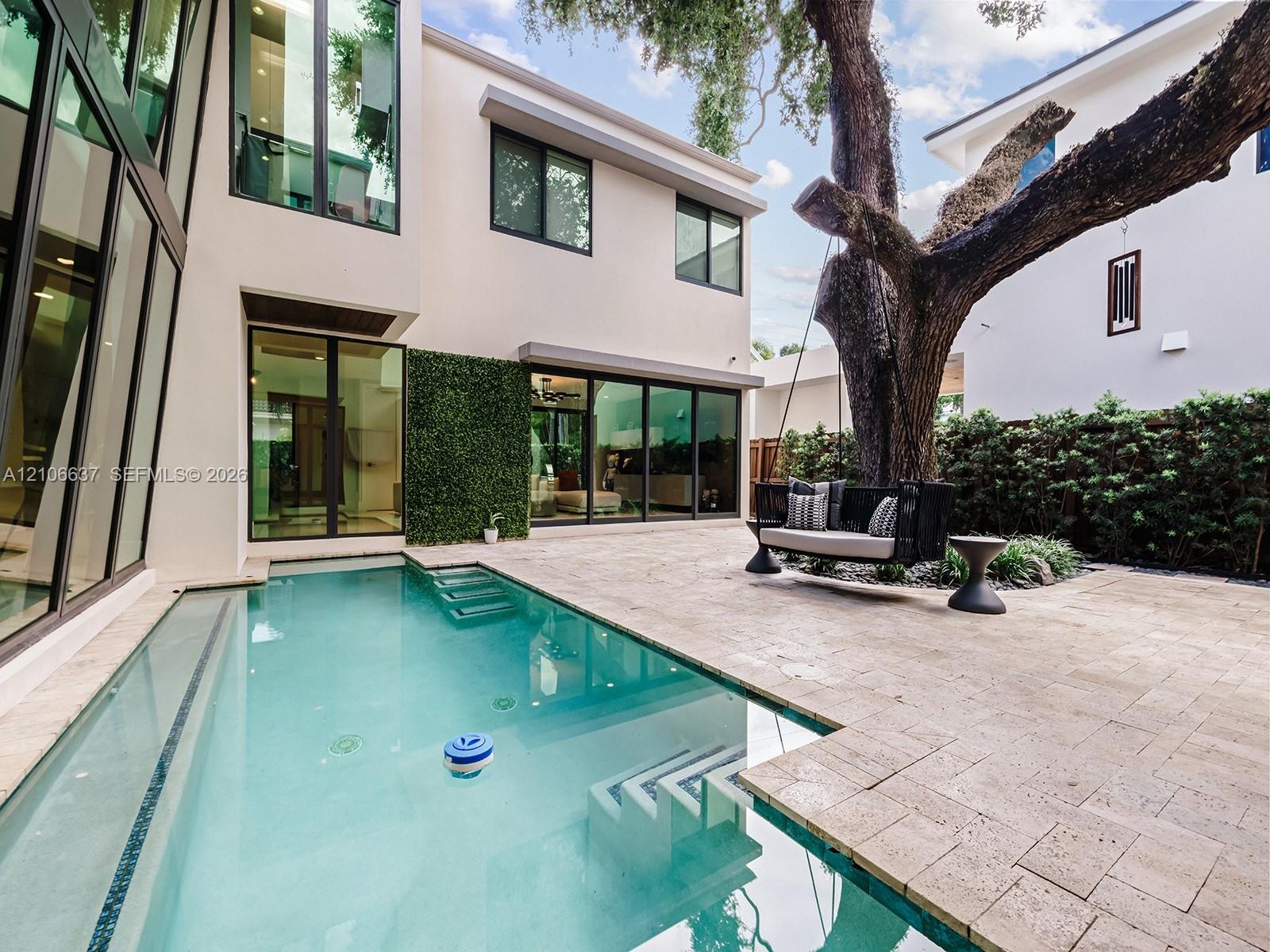Exterior courtyard featuring an in-ground swimming pool with integrated steps, stone-tile decking, full-height floor-to-ceiling windows, and mature tr