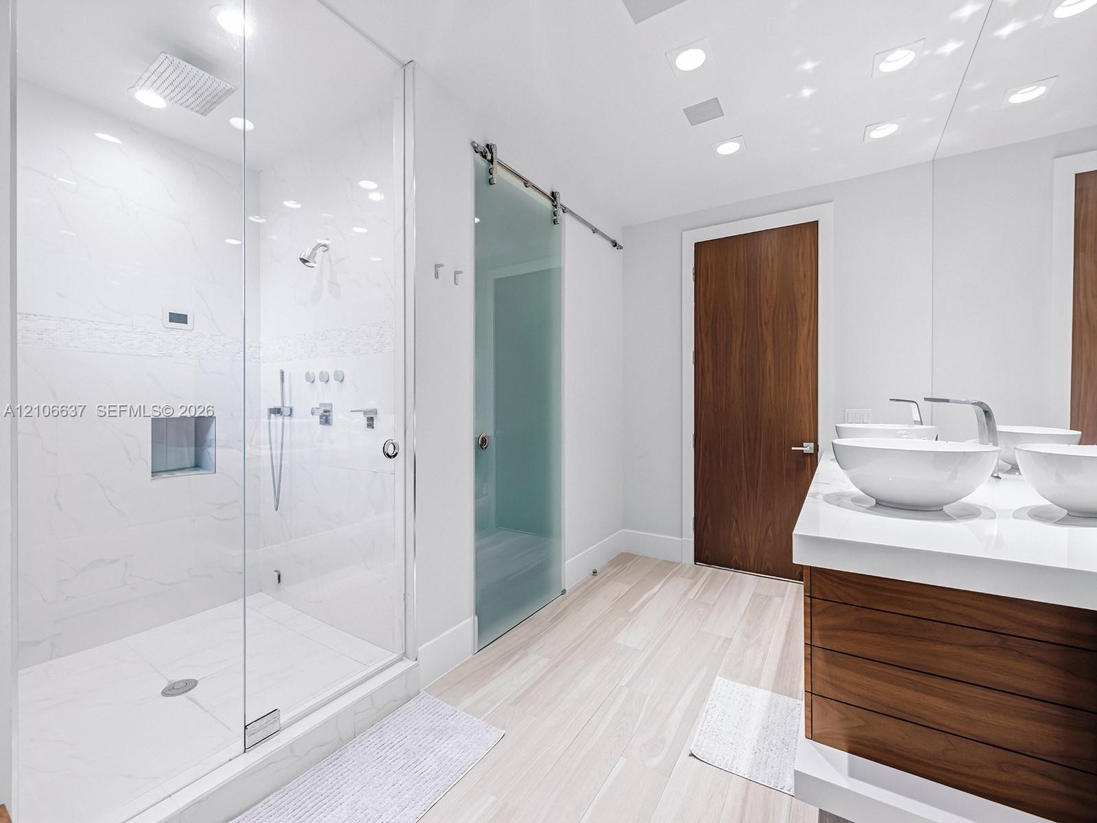 Walk-in glass-enclosed shower with wall-mounted rainfall fixture and recessed niche, dual vessel sinks atop a wood-finish vanity, polished white solid