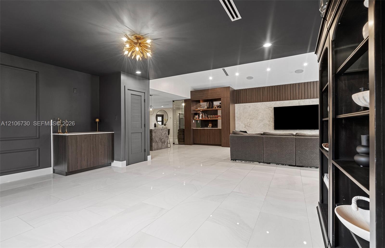 Large-format porcelain tile flooring, dark-toned wood cabinetry with integrated wet bar, textured stone accent wall, recessed lighting, and contempora