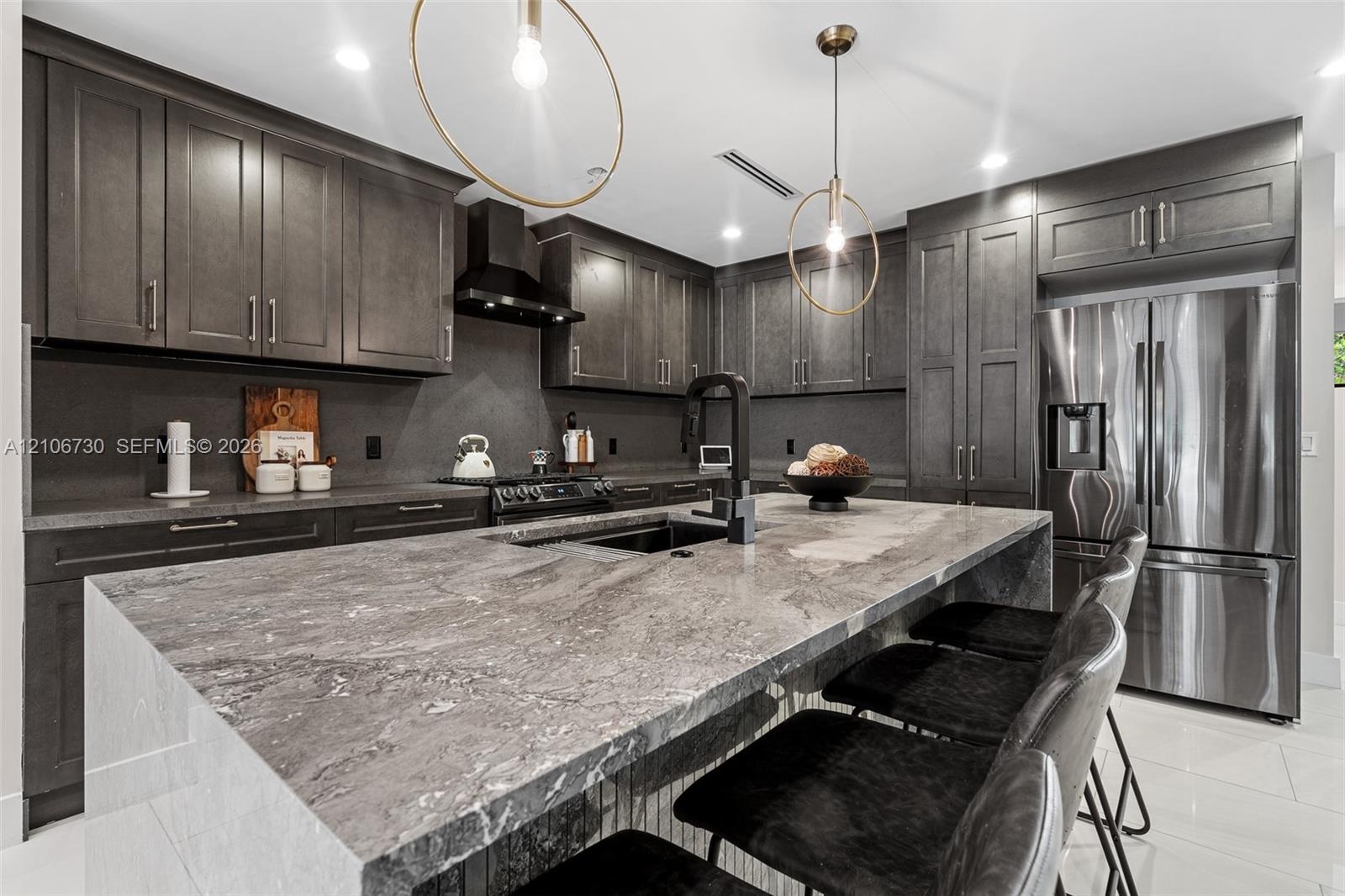 Kitchen featuring dark custom cabinetry, a stone-topped waterfall island, stainless steel appliances, a black range hood, and modern pendant lighting