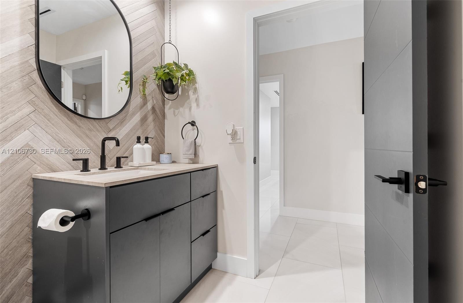 Modern vanity featuring charcoal cabinetry, a light-toned stone countertop, matte black fixtures, an arched wall mirror, and a herringbone-patterned t