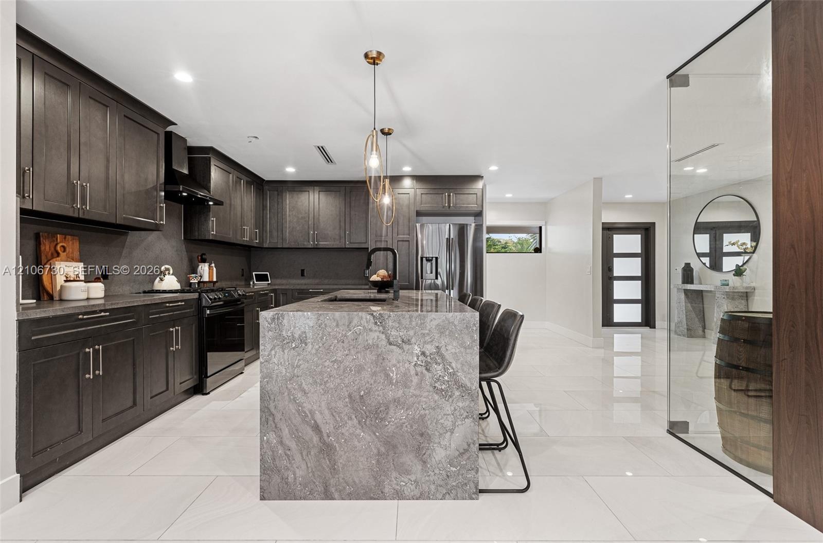 Kitchen featuring dark cabinetry, expansive stone-topped island, large-format tile flooring, stainless steel appliances, and glass-enclosed display ar