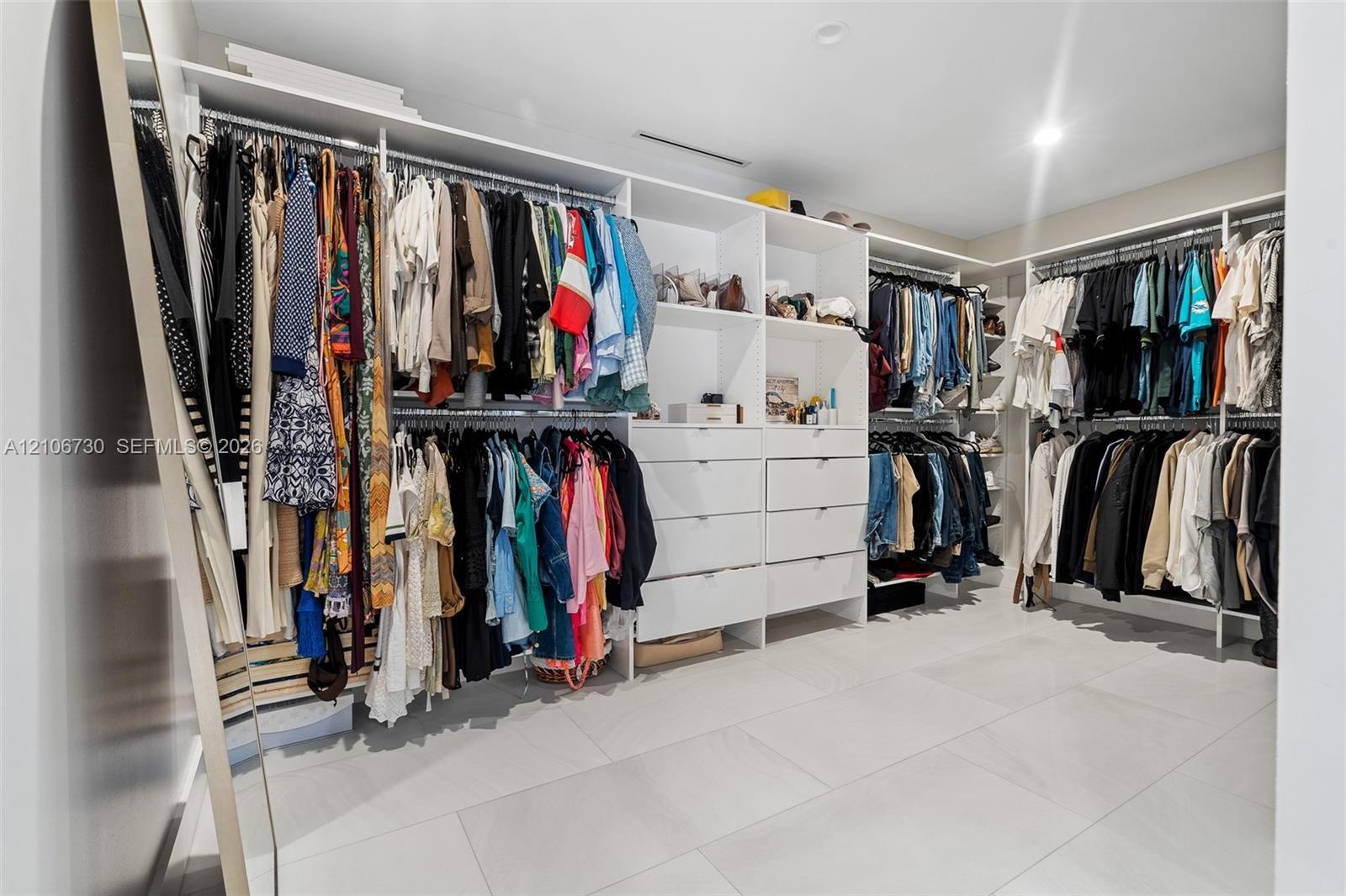 Walk-in closet featuring extensive built-in white shelving, integrated drawers, full-length hanging rods, and large-format porcelain tile flooring
