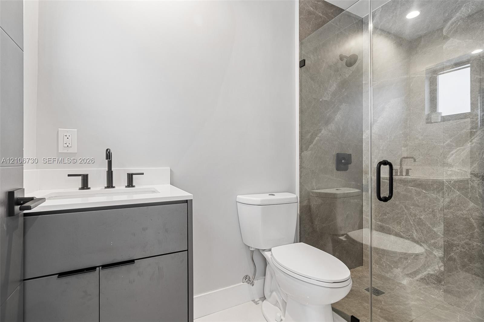 Modern vanity with a light-toned countertop, walk-in shower featuring full-height stone-finish wall tile, frameless glass enclosure, and matte black f