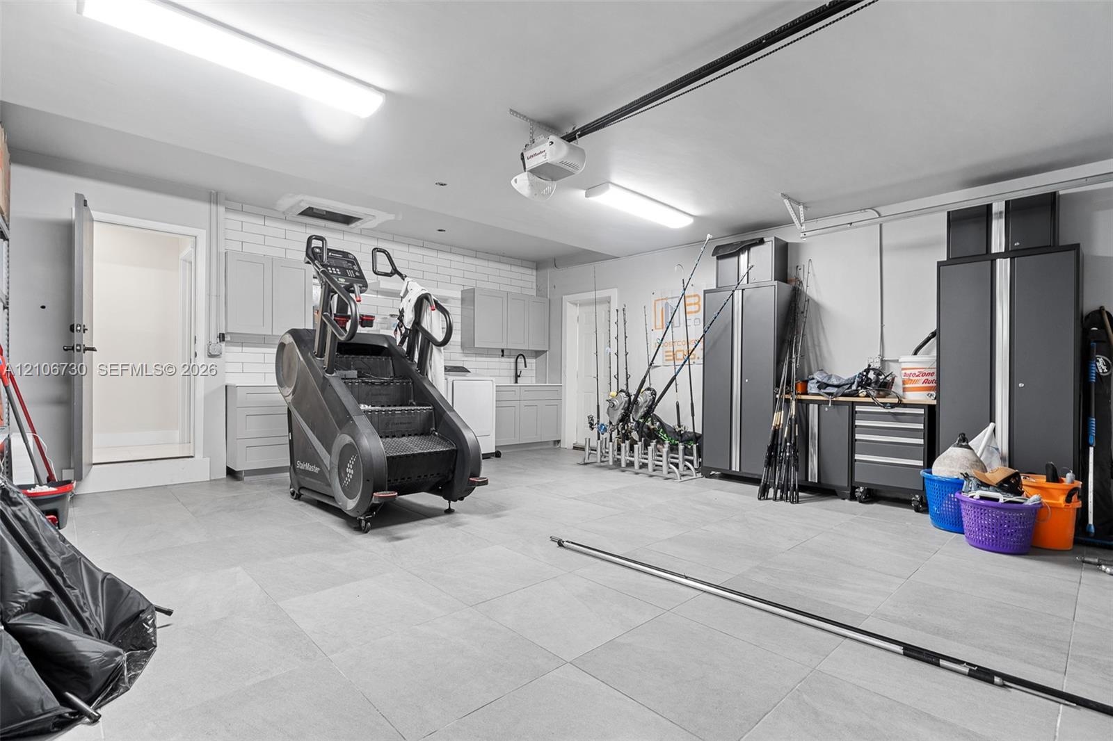Garage featuring large-format tile flooring, built-in light grey cabinetry, a subway tile backsplash, and overhead fluorescent lighting