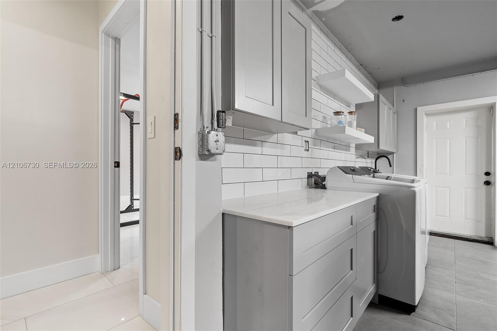 Laundry space featuring shaker-style grey cabinetry, white solid-surface countertop, white subway tile backsplash, and floating wall shelves