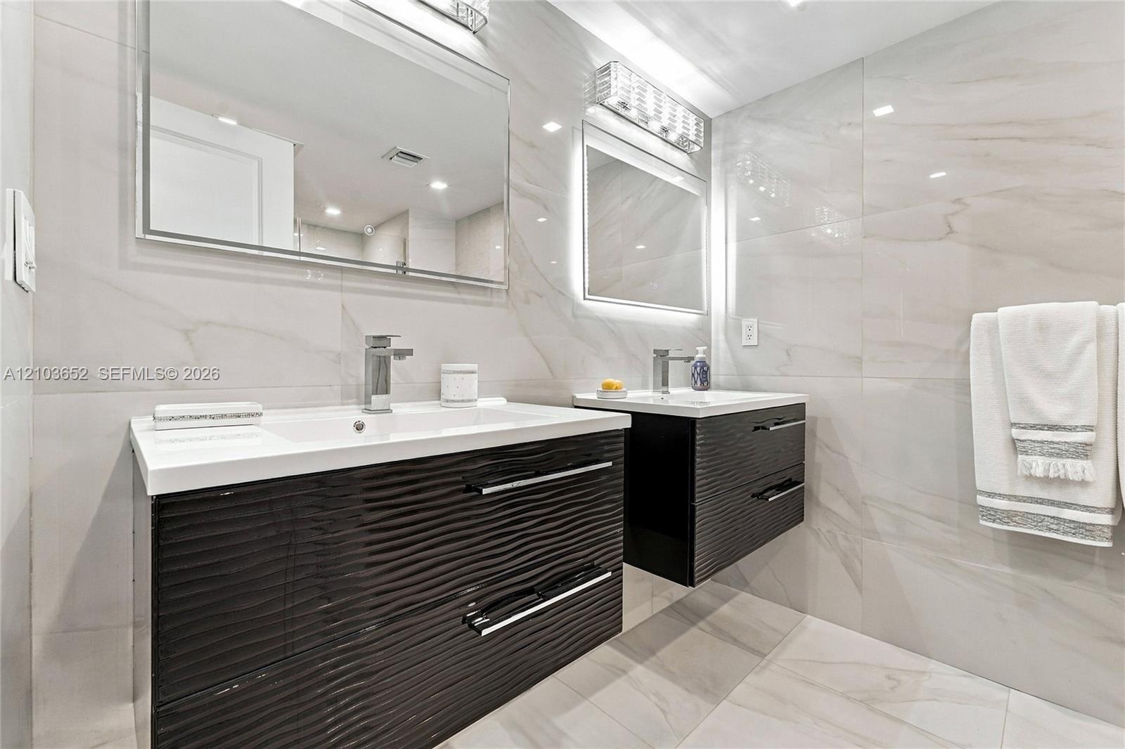 Dual floating vanities featuring dark-toned textured cabinetry, white integrated sinks, polished chrome fixtures, and full-wall porcelain tiling