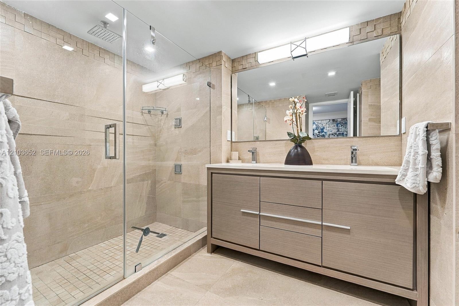 Bathroom featuring a walk-in glass-enclosed shower with a rainfall showerhead, wood-finish vanity with dual integrated sinks, and neutral stone-style 