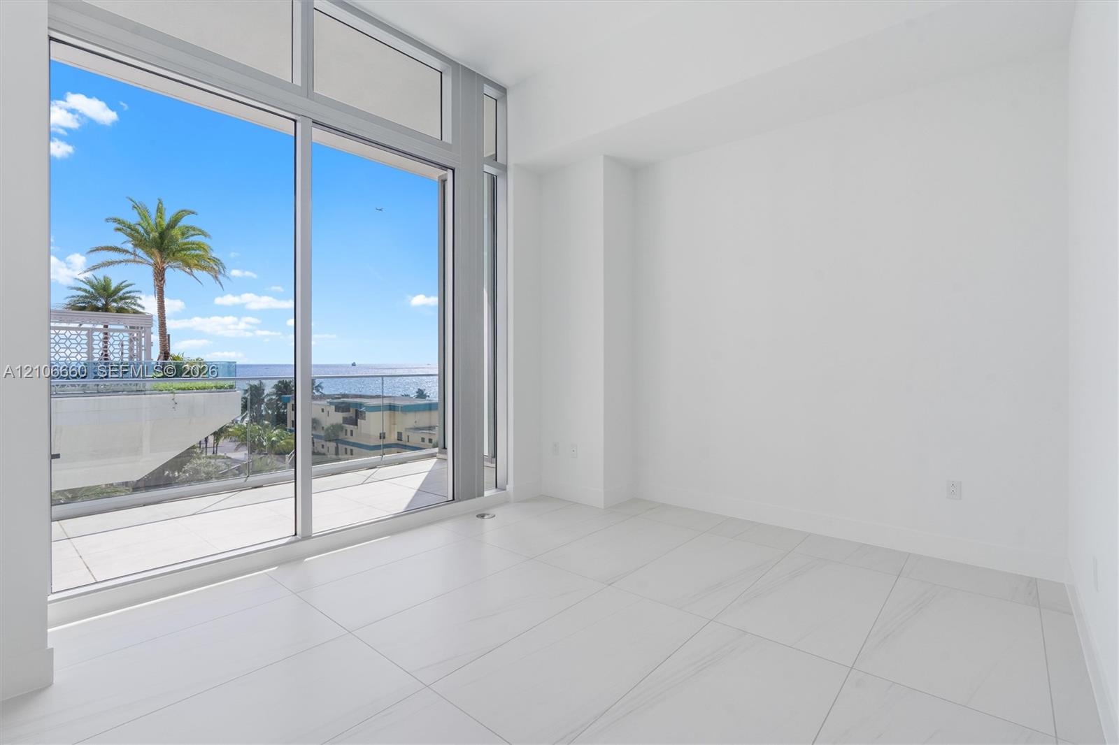 Floor-to-ceiling glass wall, large-format tile flooring, and sliding glass door opening to a private balcony with glass railing and water backdrop