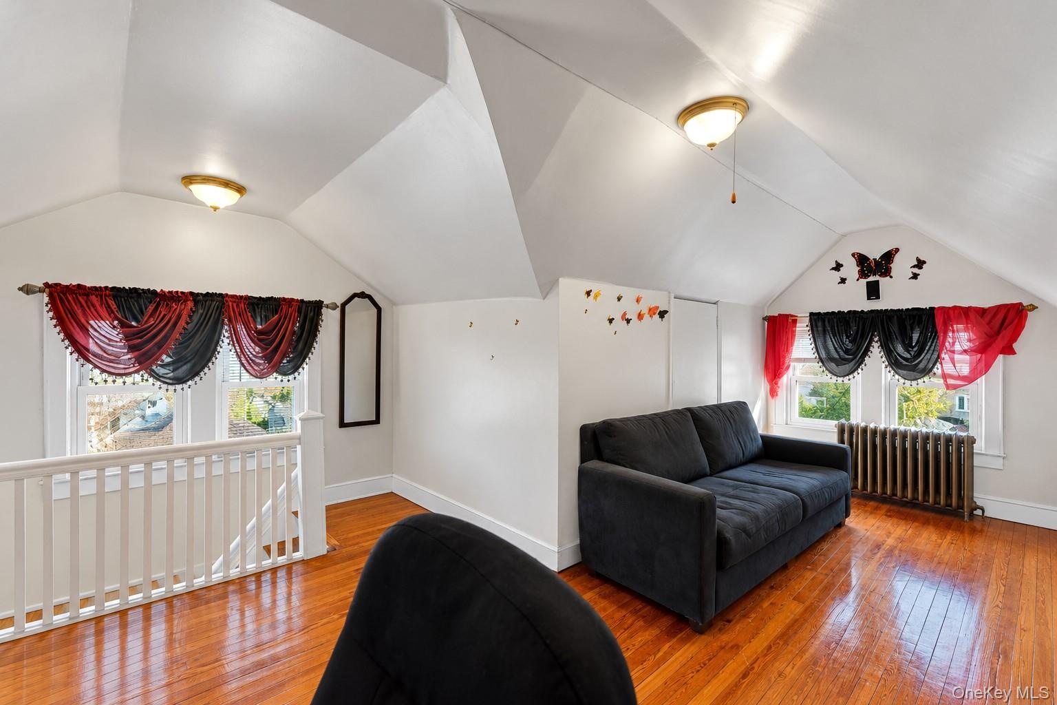 Vaulted ceiling space featuring wood-finish flooring, a metal radiator, and a white wooden staircase railing