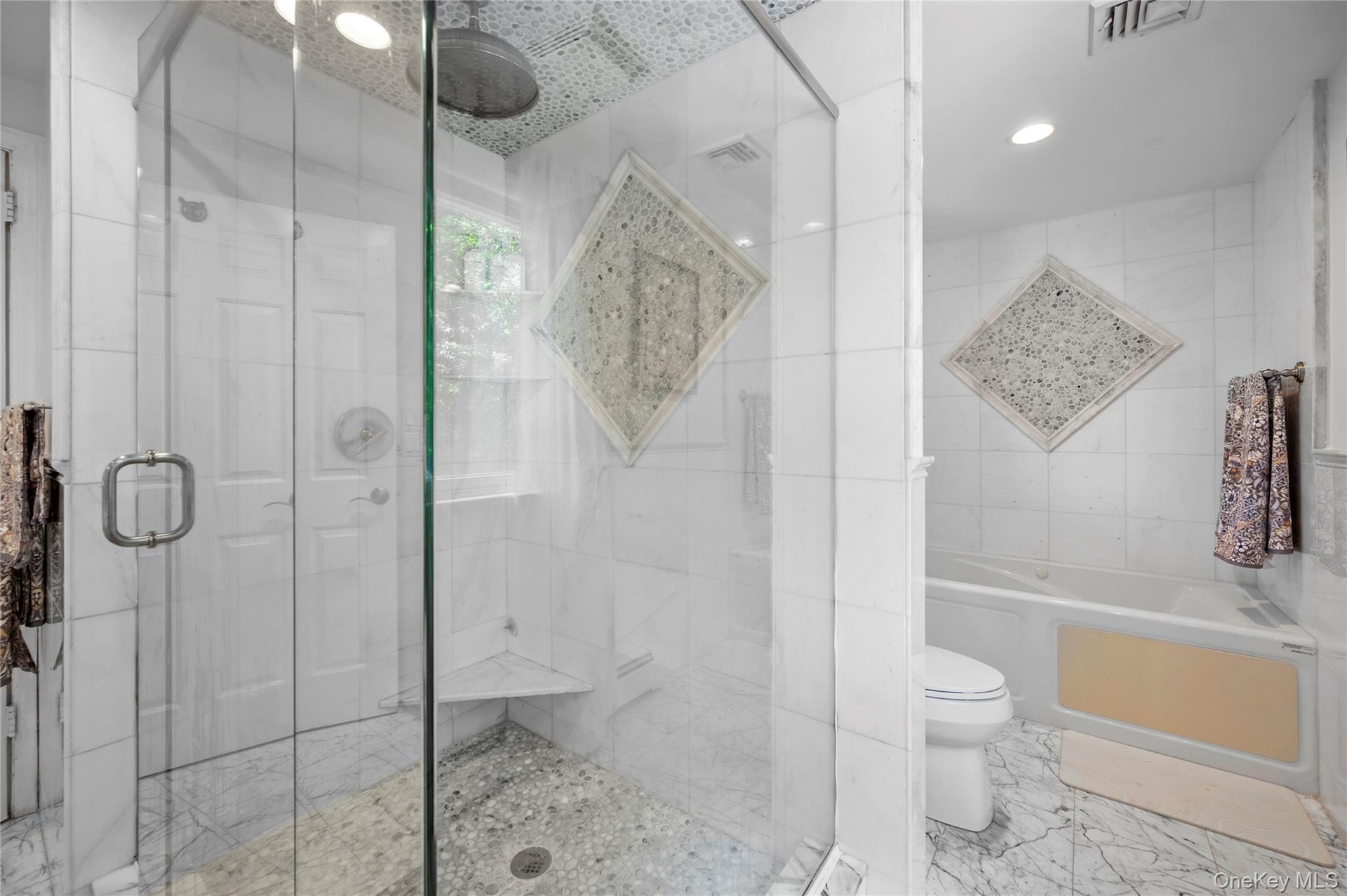 Walk-in glass-enclosed shower featuring a stone-tile floor, built-in corner seat, rain-style showerhead, and decorative mosaic tile accent