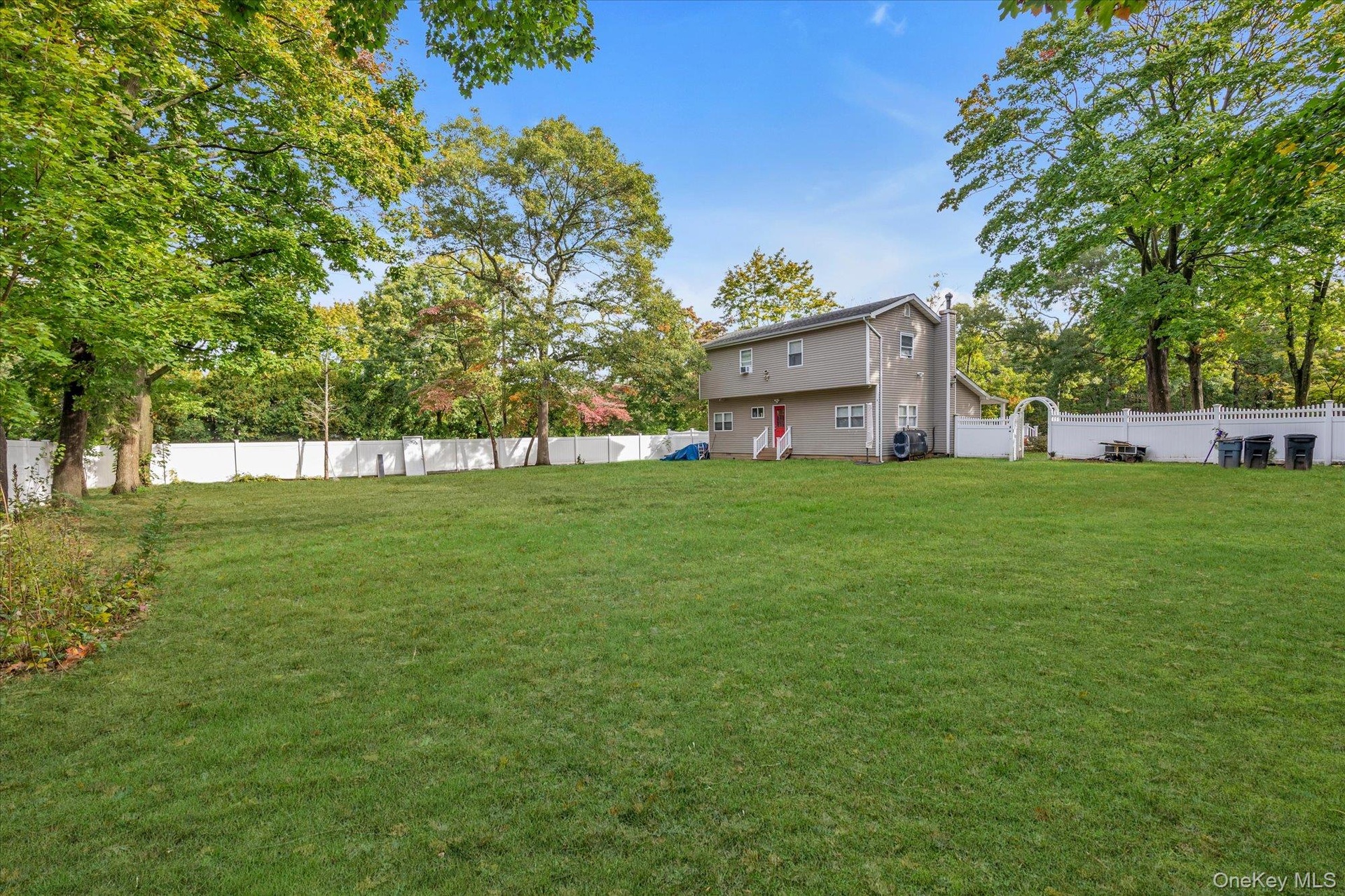 Expansive backyard featuring a large grass lawn, mature trees, and white vinyl privacy fencing