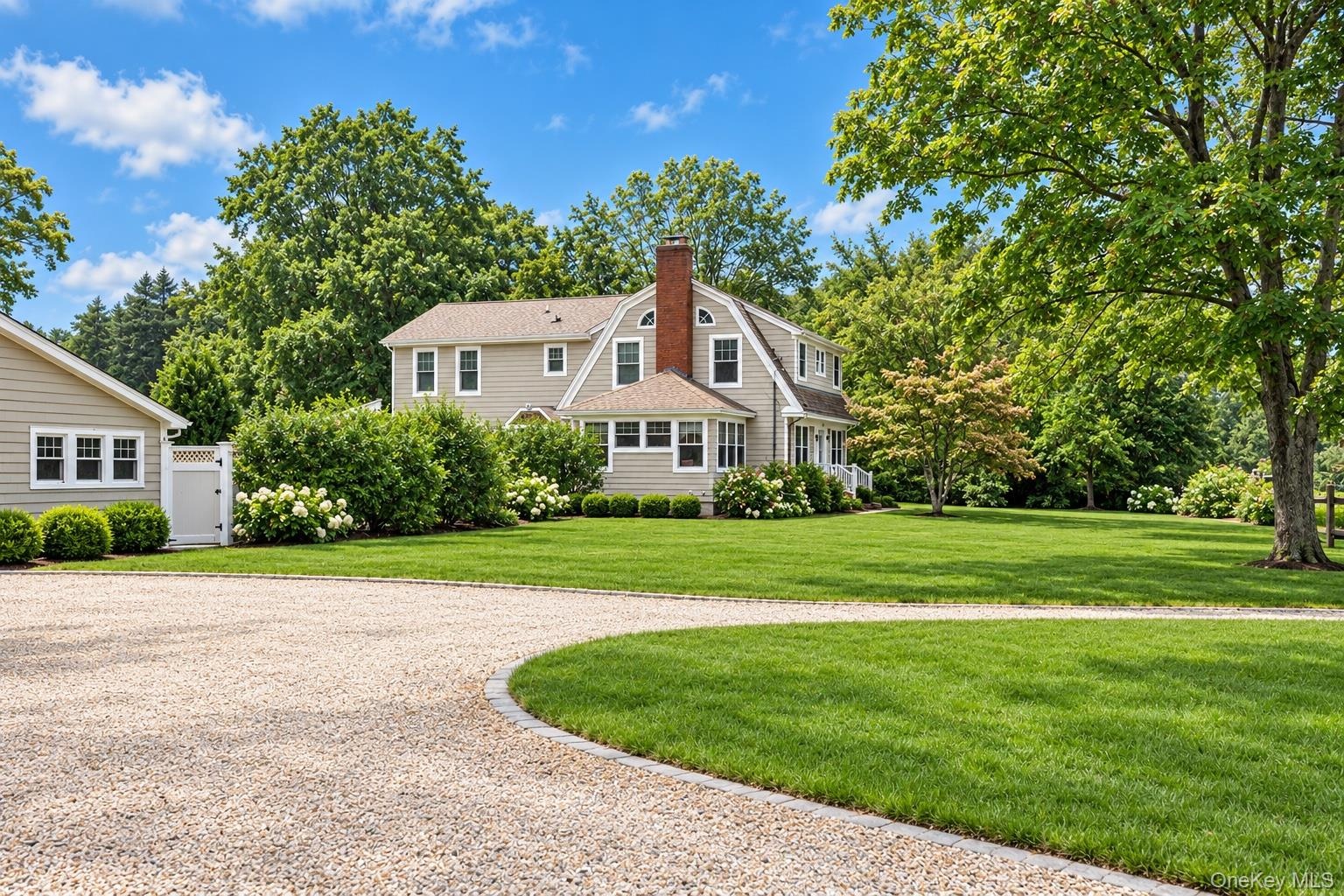 Gambrel-style exterior featuring a brick chimney, shingle siding, a sprawling lawn, and a circular gravel driveway with stone edging